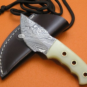 Puede incluir: Un pequeño cuchillo hecho a mano con una hoja de acero damasco y un mango de hueso blanco. El cuchillo tiene una funda de cuero con un cordón.