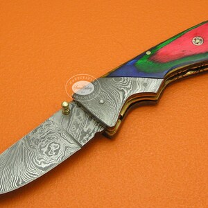 May include: A folding pocket knife with a Damascus steel blade and a colorful wood handle. The handle is red, green, and blue with a gold accent.