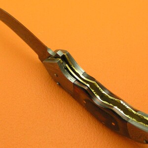 May include: A close-up of a folding knife with a curved blade. The blade is a light brown color. The handle is dark brown with gold-colored accents. The knife is set against an orange background.