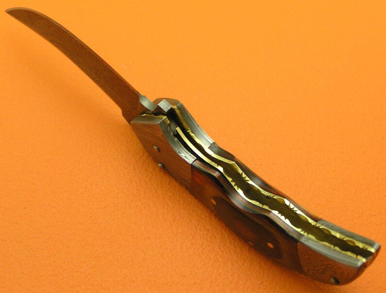 May include: A close-up of a folding knife with a curved blade. The blade is a light brown color. The handle is dark brown with gold-colored accents. The knife is set against an orange background.