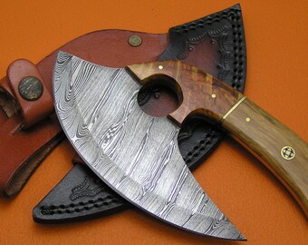 Op de afbeelding: Een Damascus stalen bijl met een houten handvat en een leren schede. De bijl heeft een gebogen blad en een gat in het handvat. De schede is bruin met zwarte stiksels.