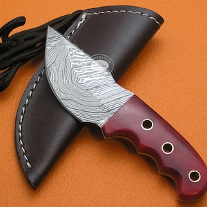 Può includere: Un piccolo coltello artigianale con una lama in acciaio Damasco e un manico in legno rosso. Il coltello ha un fodero in pelle con dettagli di cucitura decorativi. Il coltello è lungo circa 18 cm.