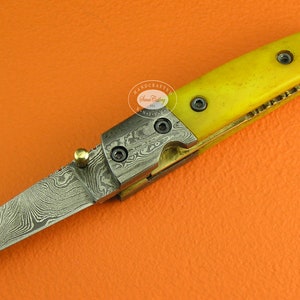 May include: A yellow-handled folding knife with a Damascus steel blade. The knife has a brass accent and a small gold ball on the handle.