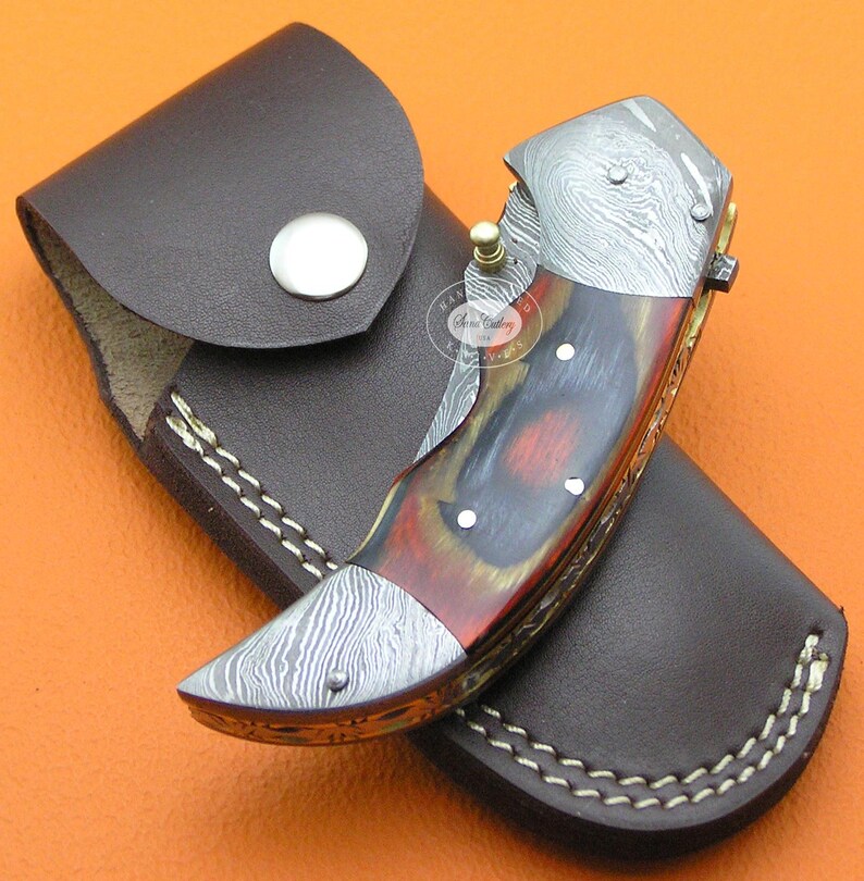 May include: A folding knife with a patterned steel blade and a handle featuring red, brown, and yellow wood grain. The knife is in a brown leather sheath with white stitching and a snap closure. The knife has a gold-colored pin.