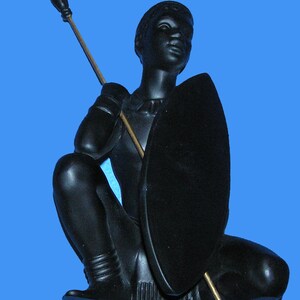 May include: A black figurine of a person kneeling, holding a spear and shield. The figure is on a blue background. The spear is held in the right hand, and the shield is held in the left hand. The figurine is detailed.