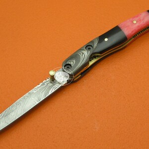 May include: A black and red folding pocket knife with a damascus steel blade. The handle is made of black and red wood with a brass accent.
