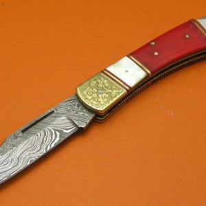 May include: A folding knife with a patterned steel blade and a red handle. The handle has white and gold accents with decorative floral patterns. The knife is set against an orange background.