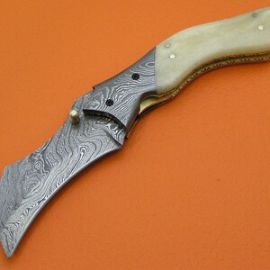 May include: A folding knife with a curved blade and a handle made of bone and patterned metal. The blade has a unique swirling pattern. The knife is set against an orange background.