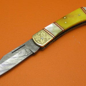 May include: A folding knife with a patterned steel blade and a yellow handle. The handle is accented with gold-colored metal and white inlays. The knife is set against an orange background.