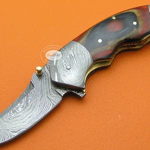 May include: A folding knife with a curved blade and a handle featuring a mix of dark red, brown, and gold tones. The blade and metal accents have a patterned, textured appearance. The knife is set against an orange background.