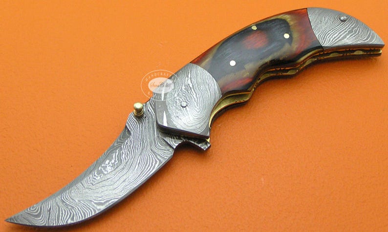 May include: A folding knife with a curved blade and a handle featuring a mix of dark red, brown, and gold tones. The blade and metal accents have a patterned, textured appearance. The knife is set against an orange background.