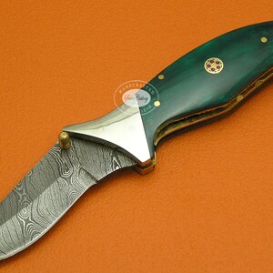 May include: A folding pocket knife with a green and gold handle and a patterned steel blade. The handle has a red and gold inlay.