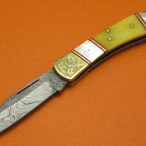 May include: A folding knife with a patterned steel blade and a yellow handle. The handle is accented with brass and mother-of-pearl inlays. The blade has a unique, wavy pattern. The knife is set against an orange background.