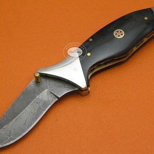 May include: A black and silver folding knife with a damascus steel blade and a black wood handle. The handle has a gold inlay with a red and gold design.