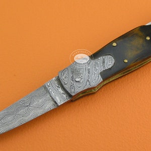 May include: A folding pocket knife with a patterned steel blade and a brown wood handle. The handle has brass accents and a brass liner.