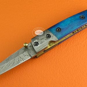 May include: A blue and gold pocket knife with a damascus steel blade. The handle is made of blue wood with a gold inlay. The knife has a small loop at the end of the handle.