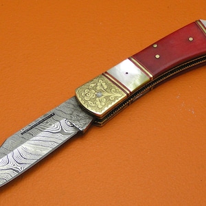 May include: A folding knife with a patterned steel blade and a handle featuring red, white, and gold accents. The handle has a red base with white inlays and gold-colored metal detailing. The blade is open, displaying a unique wave-like pattern.
