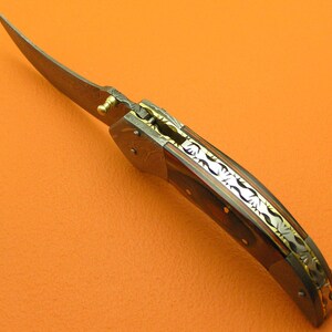 May include: A folding knife with a curved blade. The handle features a dark wood grain pattern with decorative metal accents. The blade has a unique pattern, and the knife appears to be partially open.