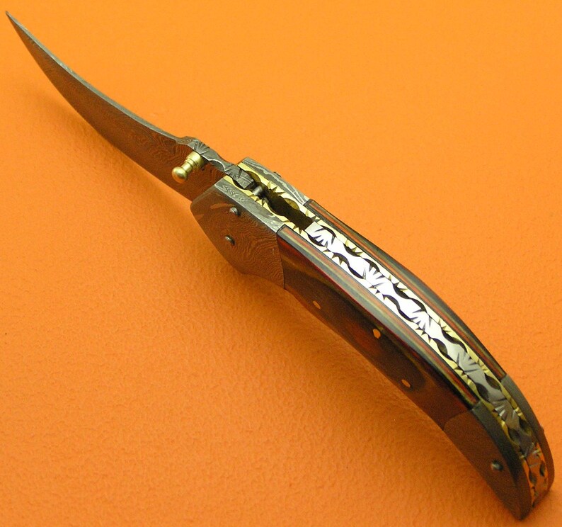 May include: A folding knife with a curved blade. The handle features a dark wood grain pattern with decorative metal accents. The blade has a unique pattern, and the knife appears to be partially open.