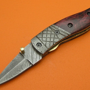 May include: A folding knife with a patterned steel blade and a dark red wooden handle. The handle is secured with black screws and features a brass accent. The blade is open, revealing its sharp edge. The knife is set against an orange background.