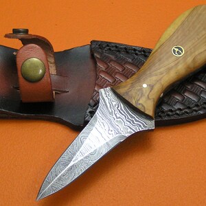 May include: A handcrafted dagger with a patterned steel blade and a wooden handle. The knife rests in a brown leather sheath with a decorative woven pattern. The handle has a small gold emblem.