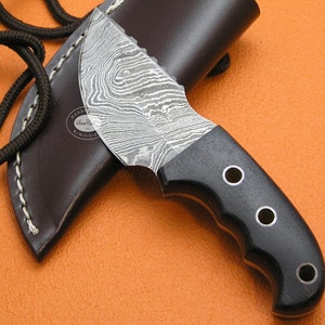 Puede incluir: Un cuchillo con mango negro, hoja de acero Damasco y funda de cuero. El cuchillo tiene un agujero para cordón y tres agujeros en el mango. La hoja es curva y tiene un borde dentado.