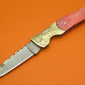 May include: A folding knife with a serrated blade and a decorative handle. The blade has a unique pattern, and the handle features a red and gold design with floral engravings. The knife is displayed on an orange surface.
