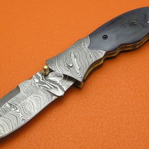 May include: A folding pocket knife with a blue wood handle and a patterned steel blade. The blade is partially open and the knife is lying on a textured orange surface.