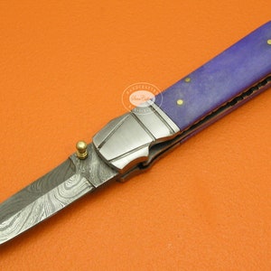 May include: A purple and silver folding knife with a decorative patterned blade and a gold accent. The handle is made of purple wood with gold accents.