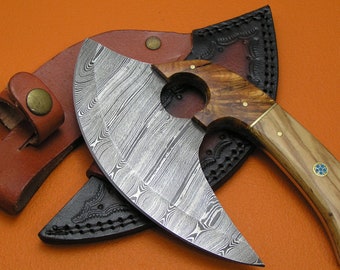 Op de afbeelding: Een handgemaakte Damascus stalen bijl met een houten handvat en een leren schede. De bijl heeft een uniek, golvend patroon op de kling en een gat in het handvat.