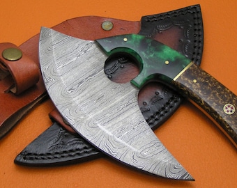 Op de afbeelding: Een Damascus stalen bijl met een groen en bruin handvat en een leren schede. De bijl heeft een uniek gebogen blad en een gat in het handvat.