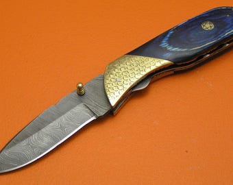 May include: A folding knife with a patterned blade and a handle featuring blue and gold accents. The handle has a brass bolster with a scale pattern. The blade has a unique pattern. The knife is set against an orange background.