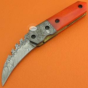 May include: A red and gold pocket knife with a curved blade and a patterned handle. The blade is made of Damascus steel and has a serrated edge. The handle is made of red bone and has a gold accent.