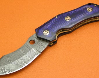 May include: A folding knife with a patterned steel blade and a purple and gold handle. The handle has gold accents and decorative rivets. The knife is open, displaying the blade against an orange background.