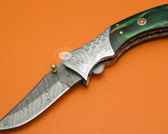 May include: A folding knife with a patterned blade and a green handle. The blade has a unique design, and the handle is accented with gold-colored rivets and a decorative emblem. The knife is set against an orange background.
