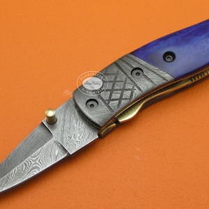 May include: A blue and silver pocket knife with a Damascus steel blade and a blue wood handle. The knife has a brass accent and a spring-loaded mechanism.