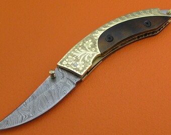 May include: A curved, gold-plated pocket knife with a wood handle and a damascus steel blade. The blade has a floral pattern etched into it.