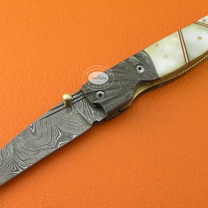 May include: A folding pocket knife with a patterned steel blade and a white mother-of-pearl handle. The handle has a decorative brass inlay.