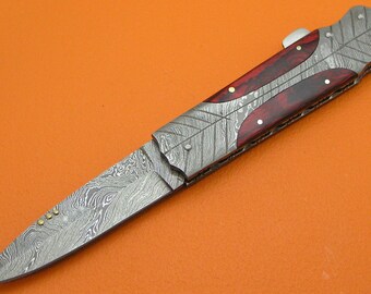 May include: A silver folding knife with a red and black wood handle. The blade is made of Damascus steel and has a decorative pattern.