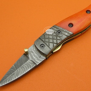 May include: A small, folding pocket knife with a Damascus steel blade and an orange bone handle. The blade has a decorative pattern and the handle has a brass pin.