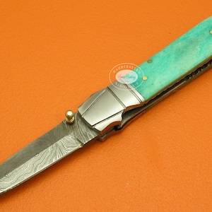 May include: A turquoise and silver folding knife with a damascus steel blade and a decorative handle. The knife has a gold-colored accent on the handle and a gold-colored loop at the end of the handle.