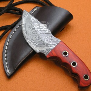 Puede incluir: Un pequeño cuchillo artesanal con una hoja de acero Damasco y un mango de madera roja. El cuchillo tiene una funda de cuero con una costura decorativa.