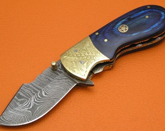 May include: A folding knife with a patterned steel blade and a blue and gold handle. The handle features a brass bolster with ornate engravings and a blue wood grain pattern. A small brass pin secures the blade.