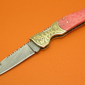 May include: A decorative folding knife with a patterned blade and a red handle. The knife features ornate gold-colored metal accents on the handle and blade base. The blade has a serrated edge near the handle.