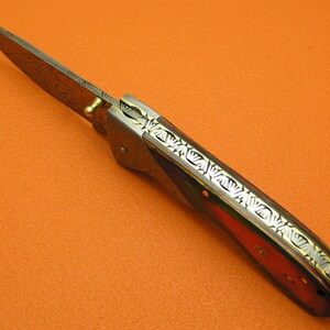 May include: A folding pocket knife with a Damascus steel blade and a wood handle. The handle has a decorative pattern of gold and silver inlay.