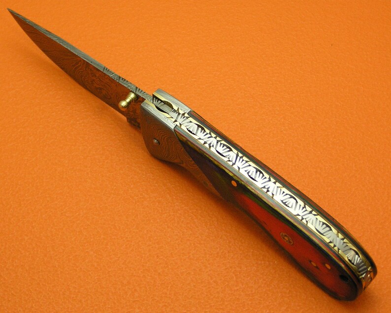 May include: A folding pocket knife with a Damascus steel blade and a wood handle. The handle has a decorative pattern of gold and silver inlay.