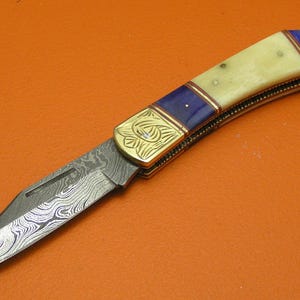 May include: A folding knife with a patterned blade and a handle featuring cream-colored material, blue accents, and gold-colored metal detailing. The knife is set against an orange background.