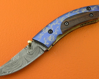 May include: A blue and gold pocket knife with a wood handle and a decorative floral pattern. The blade is made of Damascus steel.