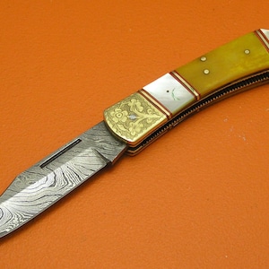 May include: A decorative folding knife with a patterned blade and a yellow handle. The handle features gold-colored metal accents and white inlays. The knife is set against an orange background.