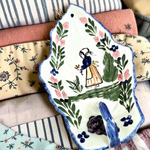 May include: A white ceramic leaf-shaped dish with a blue scalloped edge, featuring a hand-painted illustration of a person in a bonnet and apron standing in a garden, surrounded by green foliage, pink flowers, and dark blue berries.